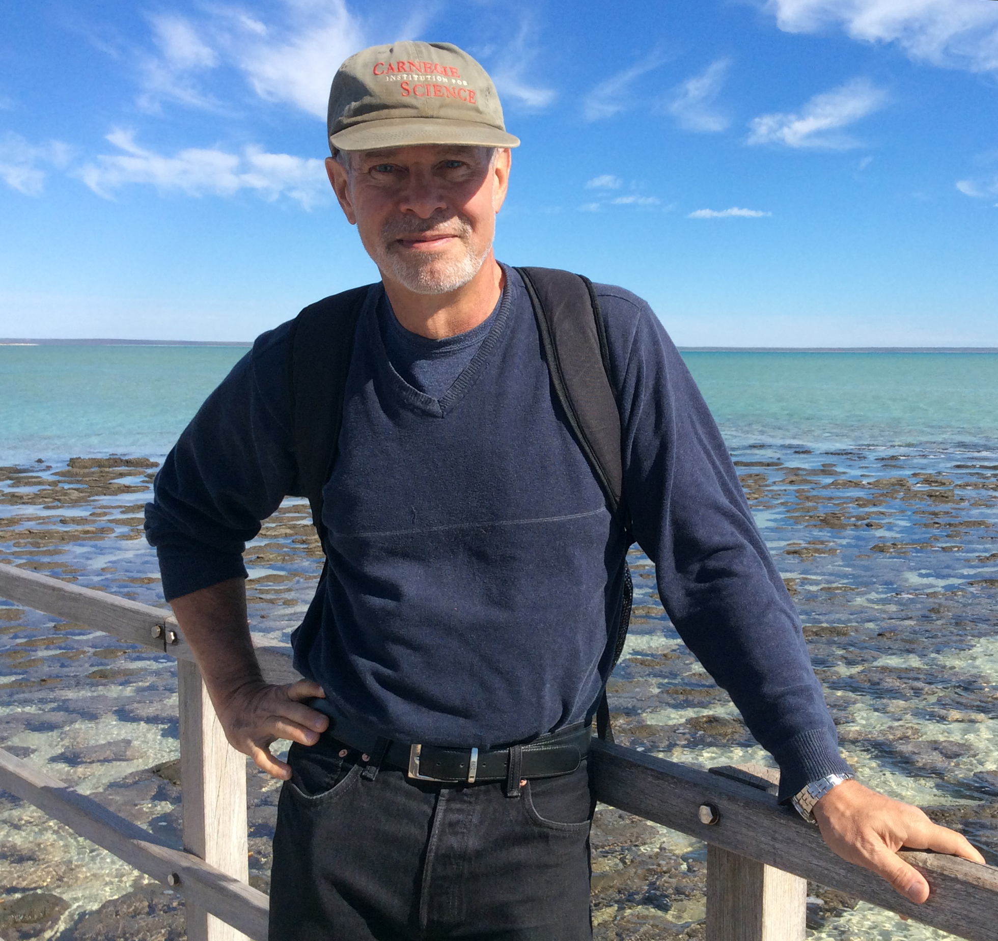 Bob Hazen on a boardwalk in front of Shark Bay, Australia, a shallow turquoise bay with dark greenish-brown stromatolite mats poking above the water surface.